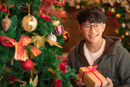 Asian man in sweater is smiling and holding gift while decorated on christmas tree with glittering balls and decorations for celebration christmas or new year in living room at homeの写真素材