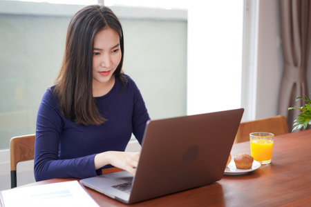 Young asian woman eating bread with a glass of orange juice while looking on laptop screen to typing business project on laptop and working at homeの写真素材
