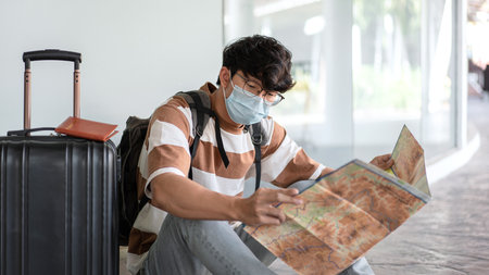 Tourist man in medical face mask is sitting on the floor with luggage during looking on the map to searching direction destination for holiday trip while going to travel with new normal lifestyleの写真素材