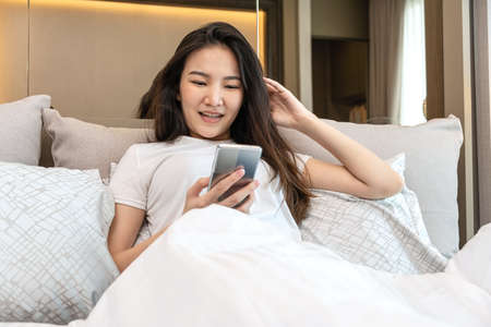 Asian woman using smartphone to chatting and watching movie on social media while lying to rest on the bed in bedroom at home and enjoy in weekendの写真素材