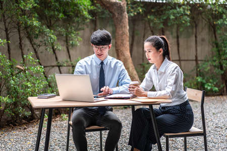 Young business woman and businessman sitting and looking about business project on laptop while holding coffee cup and talking of financial report together at outdoor officeの写真素材