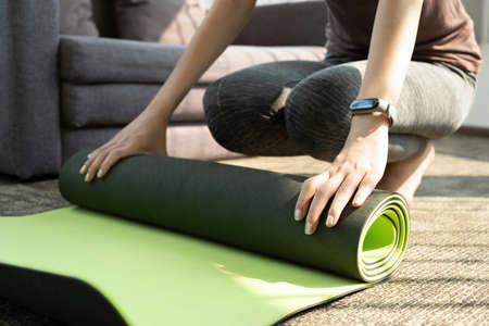 Young woman in sportswear is rolling up the mat and prepare laptop for yoga exercise class online in...