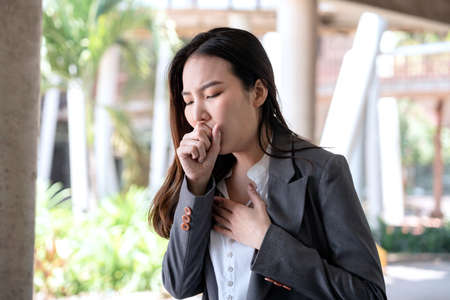 Asian businesswoman in suit, feeling sick cough and nose sneeze after work hard about new startup project while walking at outside officeの写真素材