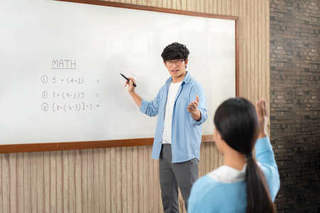 Male tutor standing in front of whiteboard is pointing to student for ask question and young students raising hands in the air to answer a question when learning in the classroomの写真素材