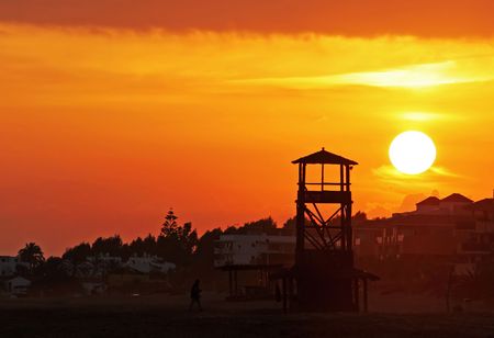 A golden orange sky and massive glowing yellow sunset at the end of the day seen from a beach in southern Spain. A silhouette from a wooden watchtower for lifeguards breaks the scene.の写真素材