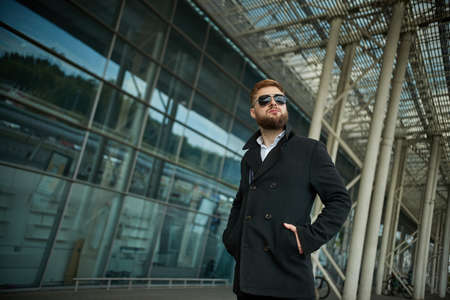 Success. Stylish young bearded man in coat, sunglasses businessman standing near the business...