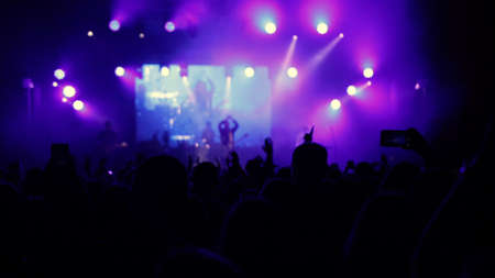 Illuminated by stage lights. Raised hands are visible. Concert crowd silhouettes in front of bright stage. People with raised hands. Concert crowd silhouettes in front of bright platform lightsの写真素材