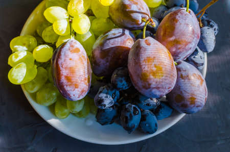 The grapes are green and black and a large plum on a white plateの写真素材