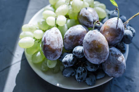 The grapes are green and black and a large plum on a white plateの写真素材