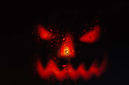 Scary face pumpkin through the wet glass with drops after rain