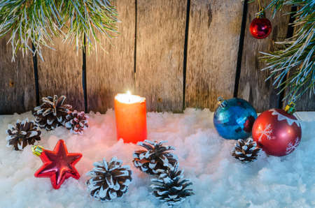 Christmas ornament, red burning candle and toys in the snow on a wooden background and tree branchesの写真素材
