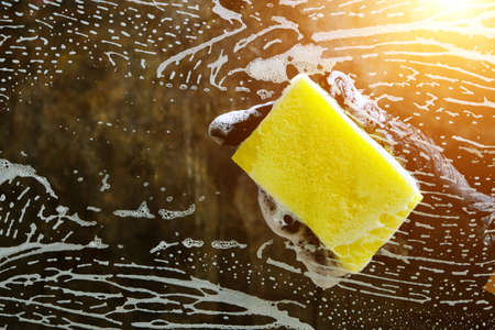 Hand in a black glove washes the glass with a foam sponge with foam on a dark background with light.の写真素材