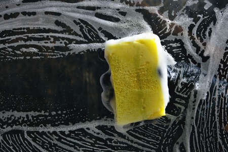 Hand in a black glove washes the glass with a foam sponge with foam on a dark background.