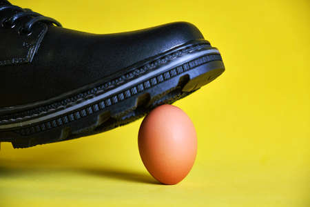Black boot on a yellow background stepped on a fragile chicken egg. Concept, weak defenselessness,...