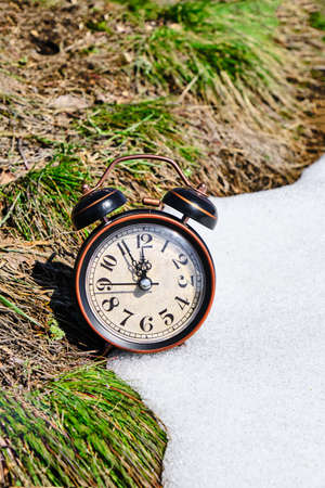 Old retro alarm clock, grass and snow, symbolizes the arrival of spring or winterの写真素材