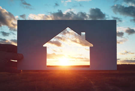 Man holds a piece of paper with a house cut out in it against the sunset. Home in nature, travel and housing. Conceptの写真素材