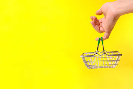 Hand holds shopping basket with its little finger, on yellow background. Concept of a trip to the supermarket.の写真素材