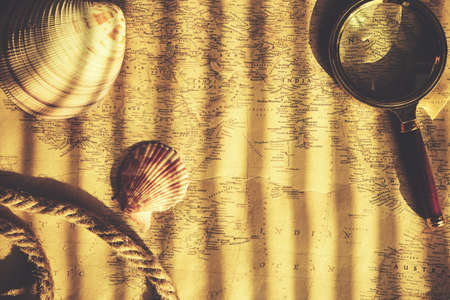 On table, an old map is pinned down with magnifying glass, shells, and rope, and sunlight shines through blindsの写真素材
