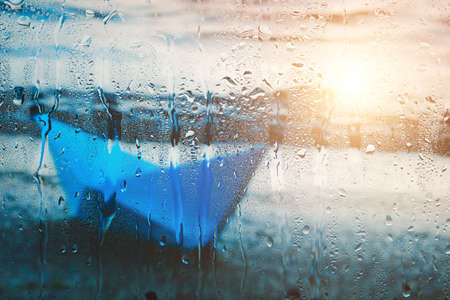 Blurry background with sunlight on water of blue paper boat washed up on beach after rain behind wet...