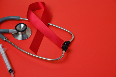 Red ribbon is symbol of AIDS, drug addiction problems, stethoscope and syringe on red background