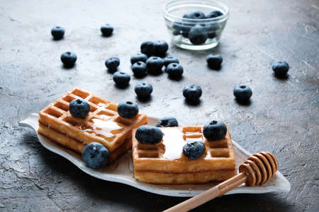 Waffles are laid out on plate, decorated with blueberries, doused with honey from honey spoon