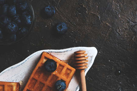 Waffles are laid out on plate, decorated with blueberries, doused with honey from honey spoon