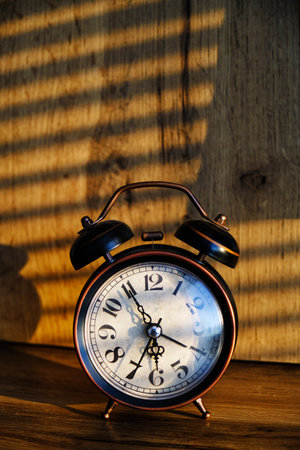 Retro alarm clock early morning on table rays sunlight through blinds in bedroomの写真素材