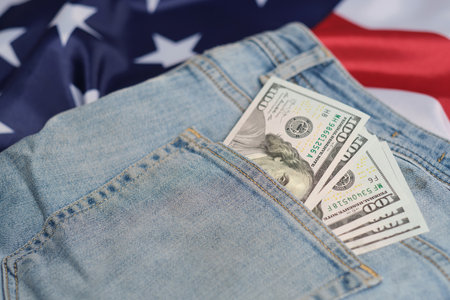 Old worn jeans are lying on flag United States America and there are hundred dollar bills in pocketの写真素材