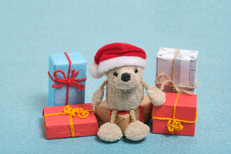 Small teddy bear in a Santa Claus hat sits on boxes with Christmas gifts.On a blue background