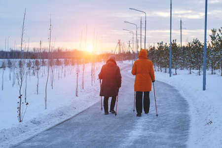 Two elderly women are engaged in sports walking in park, evening at sunset in winter