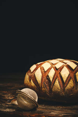 Loaf black rye bread sprinkled with flour and sieve for sprinkling on dark background