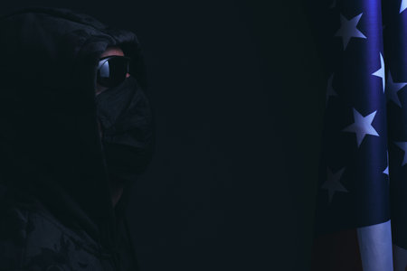 Hooded man wearing black glasses and mask looks flag of the United States America.Hacker attacker...