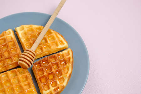 Soft muffin waffles poured with fresh honey using honey spoon, in plate on pink background