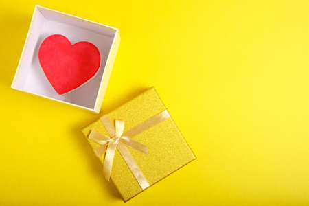 Open gift box in gold color with ribbon and bow with heart inside on yellow backgroundの写真素材