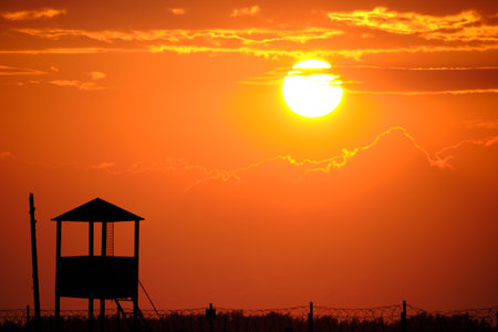 Fence with barbed wire and an old watchtower background bright sun and scarlet sunsetの写真素材