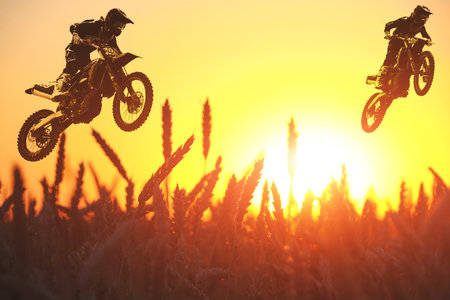 Silhouettes two motorcyclists flying over wheat field against bright sunsetの写真素材