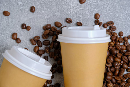 Two disposable paper cups for hot coffee lie on coffee beans on light concrete backgroundの写真素材