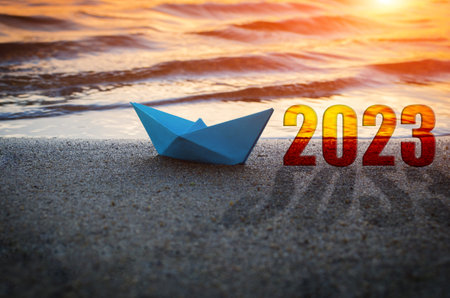 A blue paper boat and the year 2023 on a sandy beach.Against the background of sunset.The concept of...