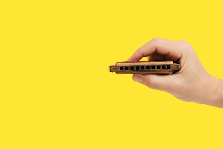 Hand Holding Harmonica on Yellow. Musical charm, perfect for nostalgic designs or music-related promotionsの写真素材