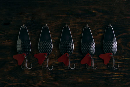 Five silver spoon lures with red tails laid out in row on dark, textured wooden background.