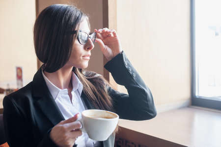 beautiful business woman sitting in a cafe near the window and drinking coffee while holding sunglasses with handの写真素材