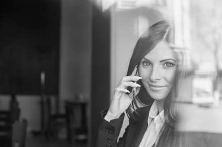 Business woman sitting in a cafe near the window smiling and talking on the phone. Black-and-white photo in the reflectionの写真素材