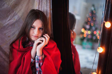 a teenage girl was hiding a red blanket, sitting near the mirror which reflects the Christmas treeの写真素材