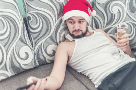 Sad young man lying on the sofa at Christmas. A lazy guy in a Christmas hat drinking champagne.の写真素材