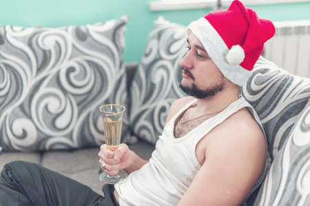 Sad young man lying on the sofa at Christmas. A lazy guy in a Christmas hat drinking champagne.の写真素材