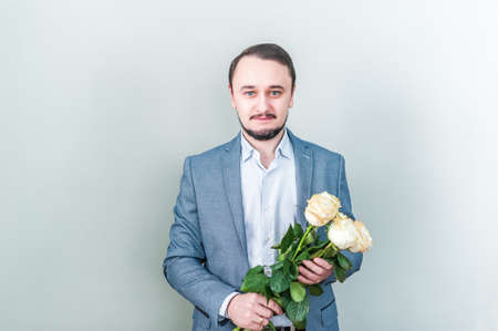 Handsome man with beard standing against a grey background with a bouquet of white roses. Business man gives roses to the occasion.の写真素材