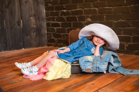 Little girl in white hat lies in the suitcase with things and looking at the camera. The child swinging his legs, smiling happily. Preparing to travel with childrenの写真素材