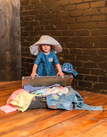 Little girl in a blue dress and white hat stands next to a suitcase with things and looking at the cameraの写真素材