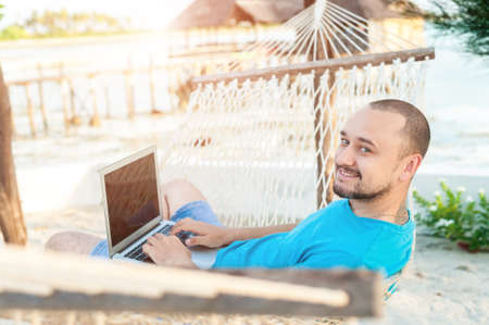 A man with a beard lying in a hammock with a laptop. Remote work on the beachの写真素材