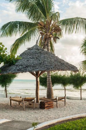Sun loungers on the white sand and an umbrella of palm leaves in the Golden glow of the tropical sun.の写真素材